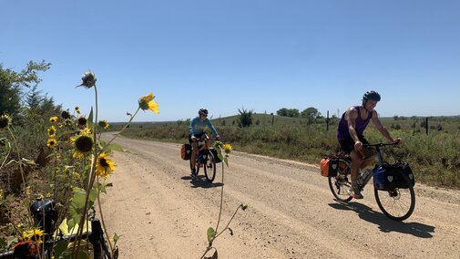 Two cyclists biking on a dirt road with sunflowers in the foreground