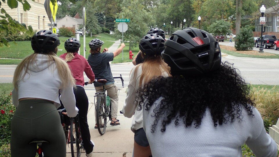 Cyclists wearing helmets pause on a neighborhood path, one pointing ahead toward a street sign.